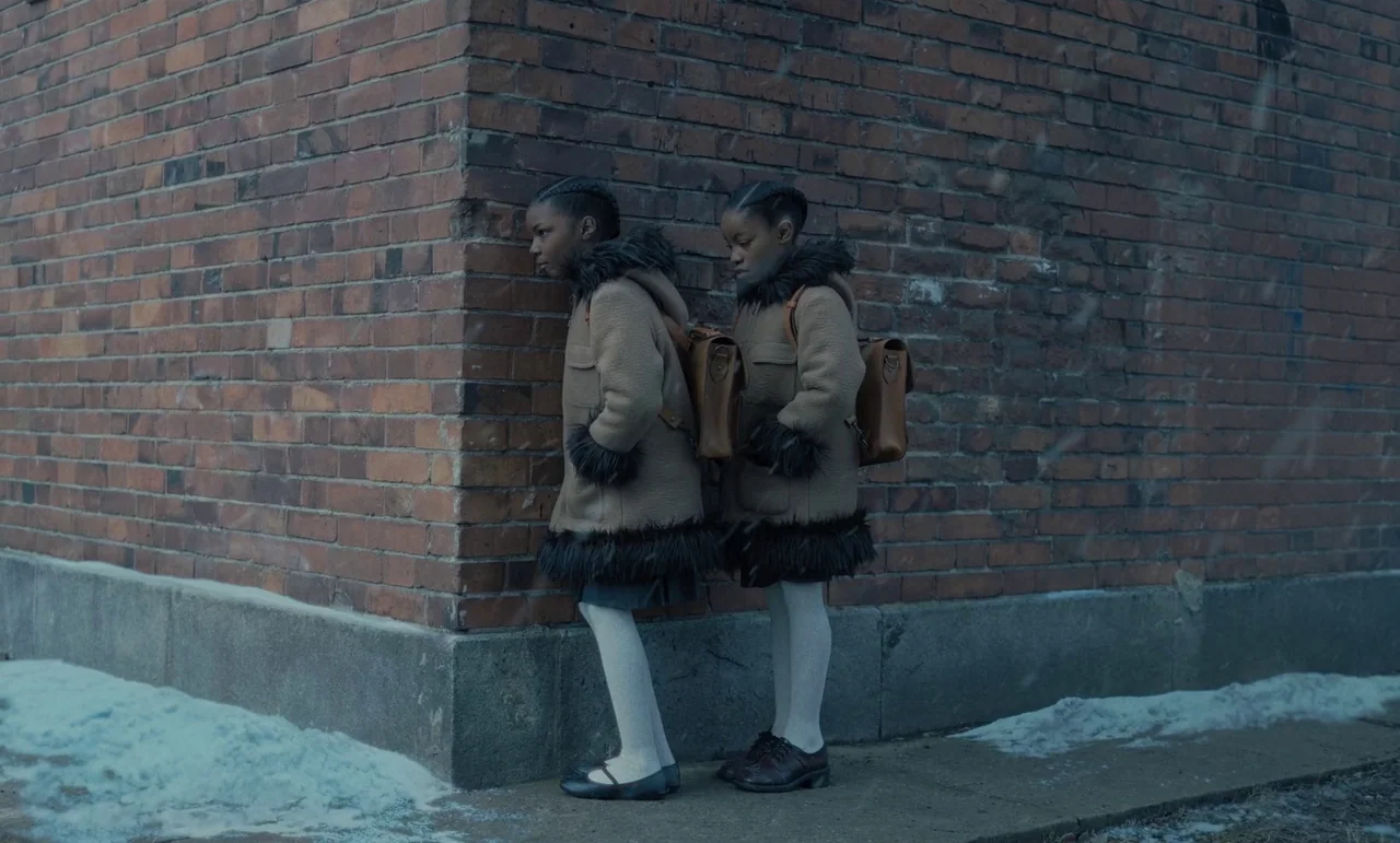 The Silent Twins (2022) movie still: fullbody — Two identical young girls stand at the corner of a red brick building. Light snow falls on the…