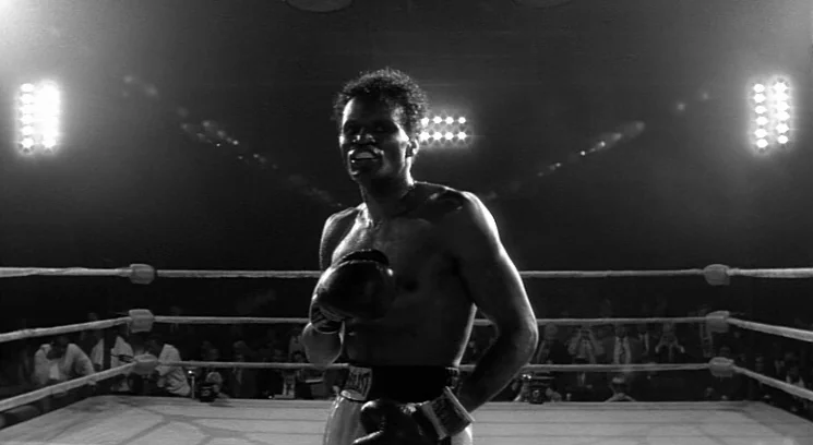 Raging Bull (1980) movie still: medium — A boxing ring is surrounded by shadowy silhouettes of a seated audience. A shirtless boxer in trunks stands…