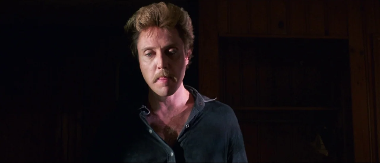 At Close Range (1986) movie still: medium — A dimly lit interior space features dark wood paneling on the wall behind. A man with a mustache stands…