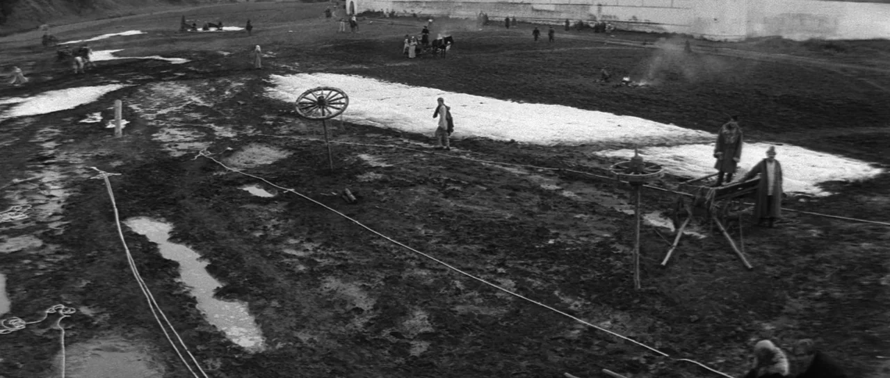 Andrei Rublev (1966) movie still: wide — A wide shot shows a vast, muddy field with patches of snow and debris. Small figures walk across the…