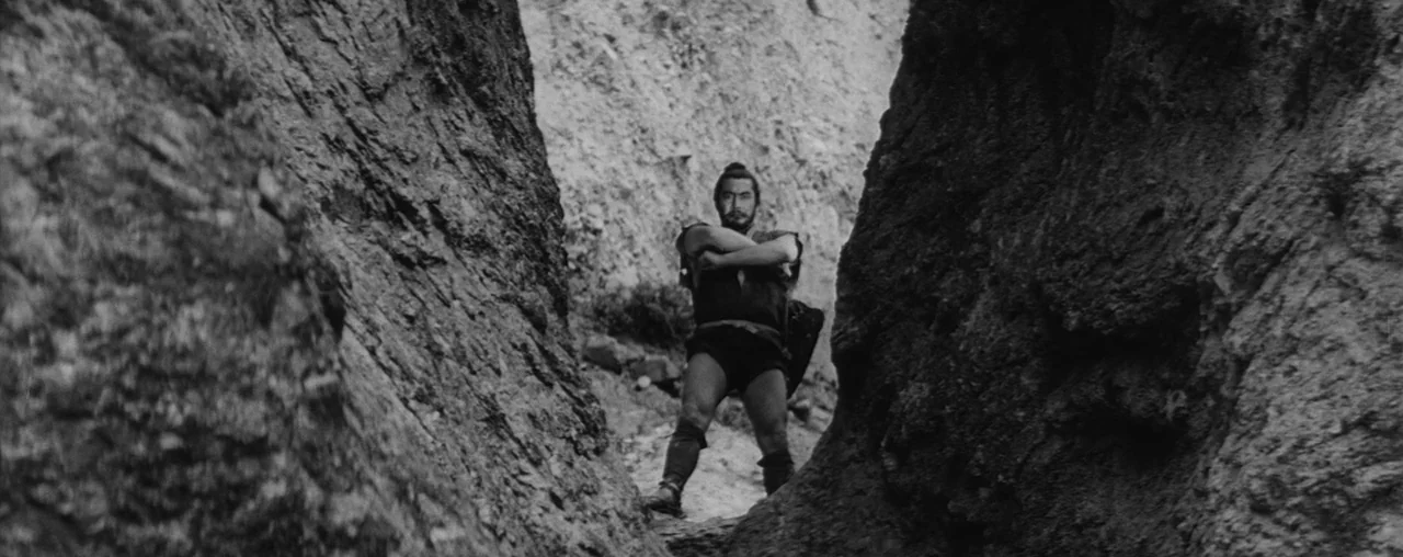 The Hidden Fortress (1958) movie still: fullbody — A narrow passage between two rough, craggy stone canyon walls. A man stands at the center of the…