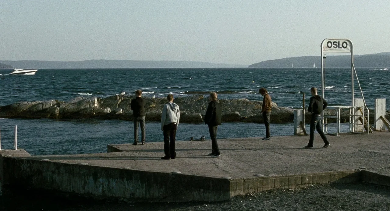 Reprise (2006) movie still: wide — A concrete pier extends into the sea under a clear bright sky. Five young men stand in a line facing the open…