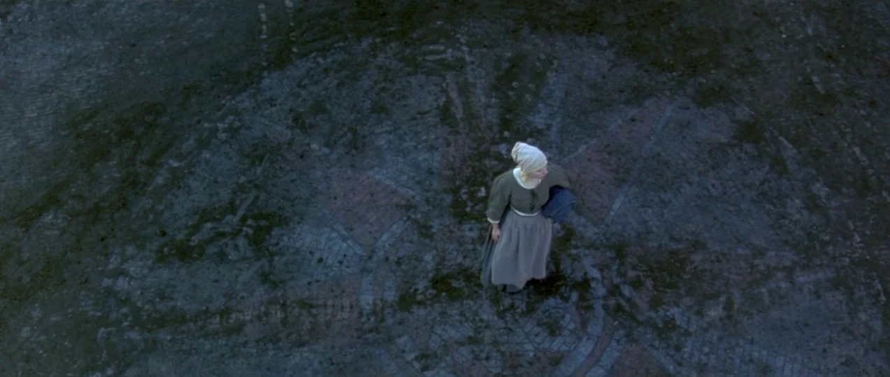 Girl with a Pearl Earring (2003) movie still: wide — A stone courtyard is paved with reddish bricks forming a large circular pattern. A woman stands…