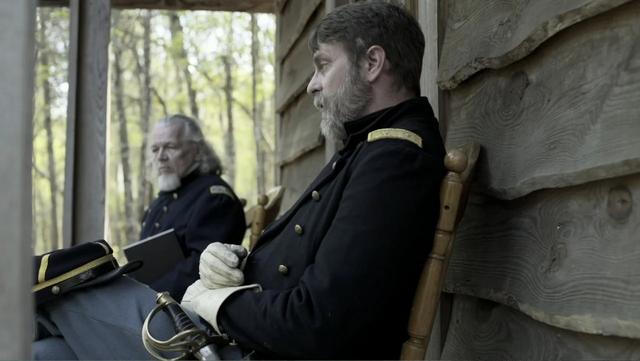 Kill Cavalry (2021) movie still: medium — Two Union officers sit on a wooden porch outside a simple log structure. They are dressed in historical…