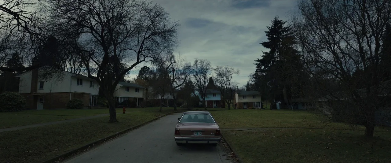 Longlegs (2024) movie still: wide — An empty suburban street is lined with mid-century houses and leafless deciduous trees. A single tan sedan…