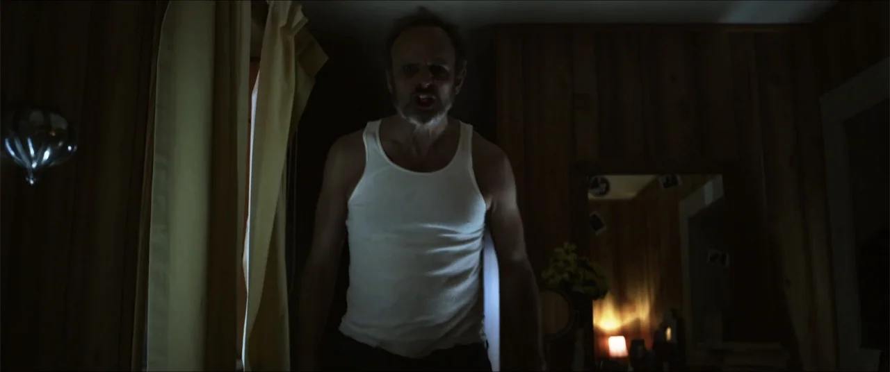 Captive (2020) movie still: medium — A dark room with wood paneled walls contains a man standing near a window. The man wears a white tank top and…