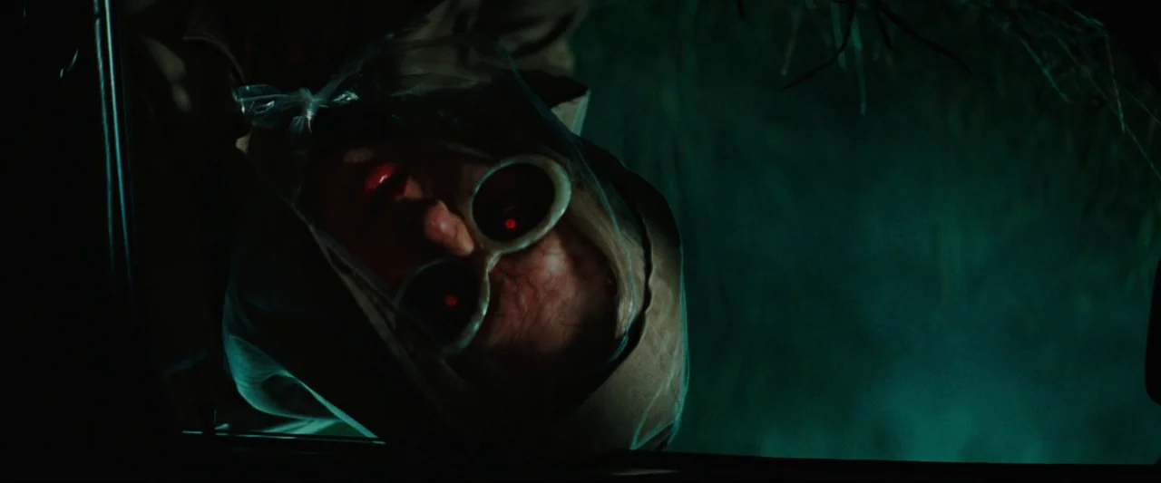 Cuckoo (2024) movie still: closeup — A dark, exterior night setting showing only a small sliver of a vehicle frame. A person wearing thick glasses…