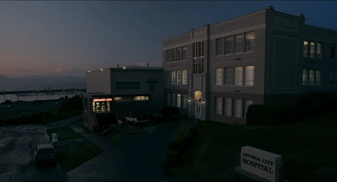 The Ring Two (2005) movie still: establishing — A multi-story hospital building is seen at dusk with lit windows and an emergency entrance. A stone…
