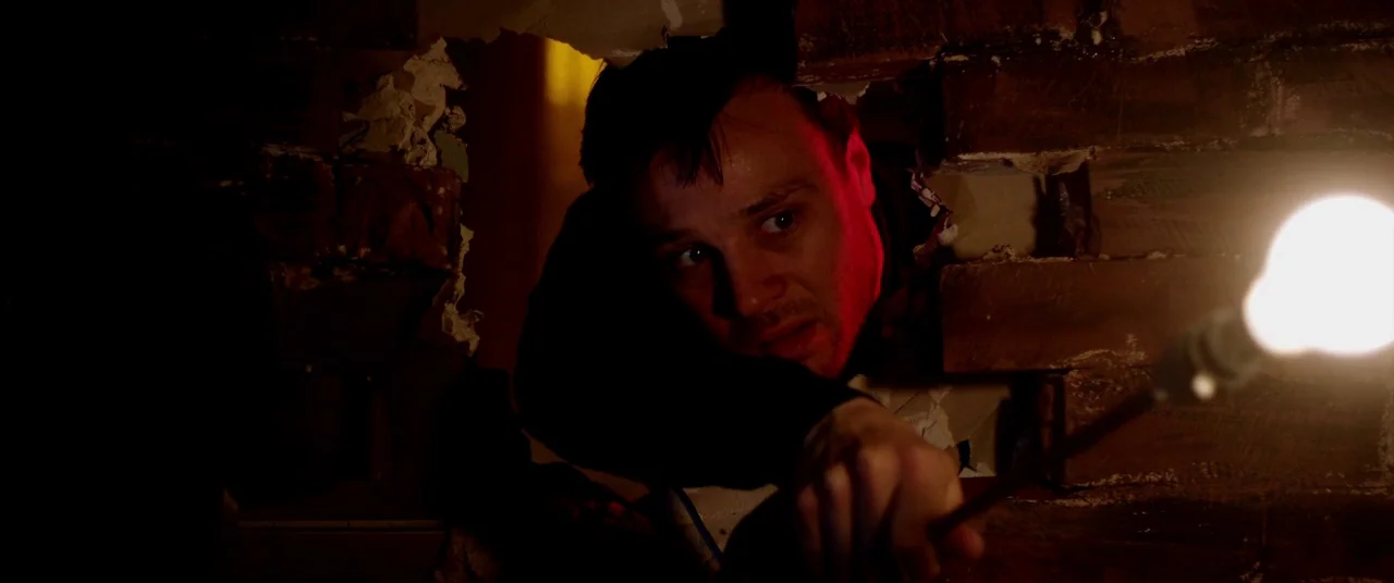 The Canal (2014) movie still: medium — A man peers through a jagged hole in a dark, weathered brick wall. He holds an exposed incandescent light…