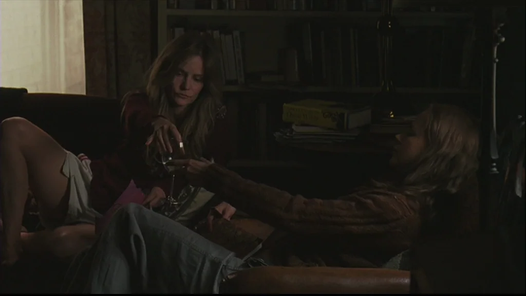 Margot At The Wedding (2007) movie still: medium — A dimly lit room with a bookshelf and a table in the background. Two women recline on a sofa…