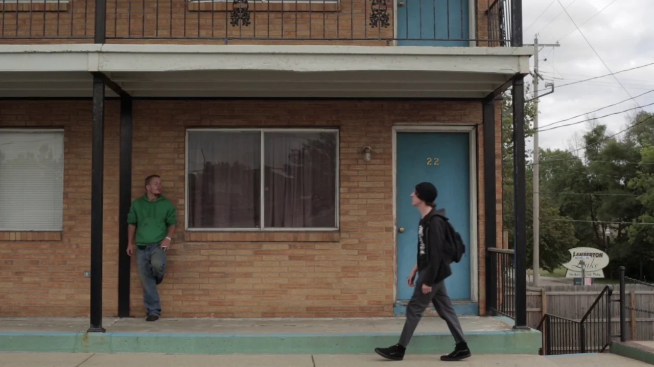 Buzzard (2014) movie still: fullbody — A two-story brick motel exterior features a blue door marked with the number 22. One man stands leaning…