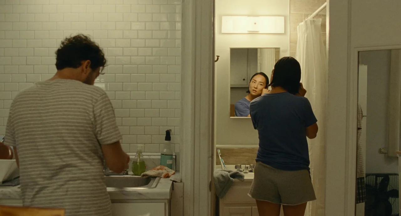 Past Lives (2023) movie still: medium — A residential bathroom features white subway tiles and a vanity mirror. A man washes his hands at the sink…