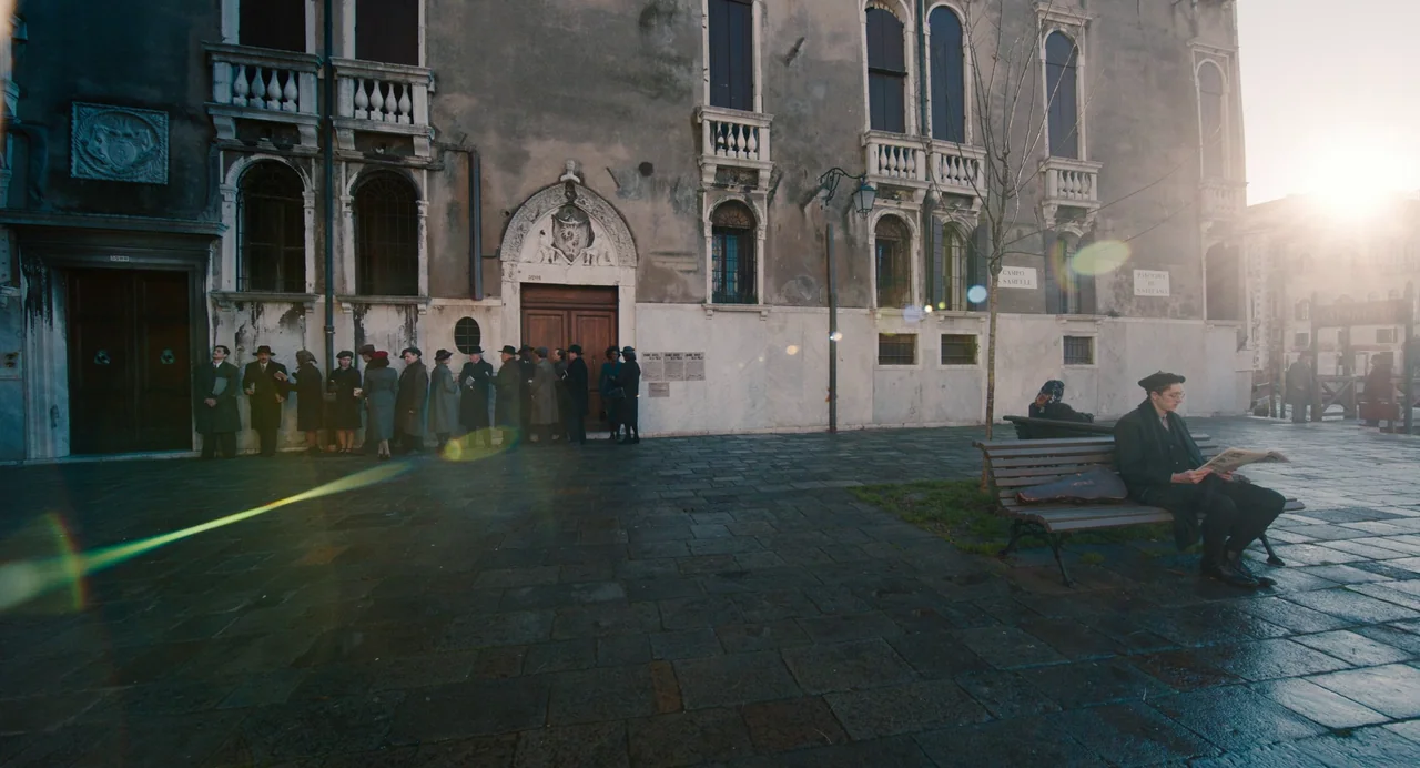 A Haunting In Venice (2023) movie still: wide — A stone paved plaza in Venice features an old building with arched stone doors. A man sits on a…