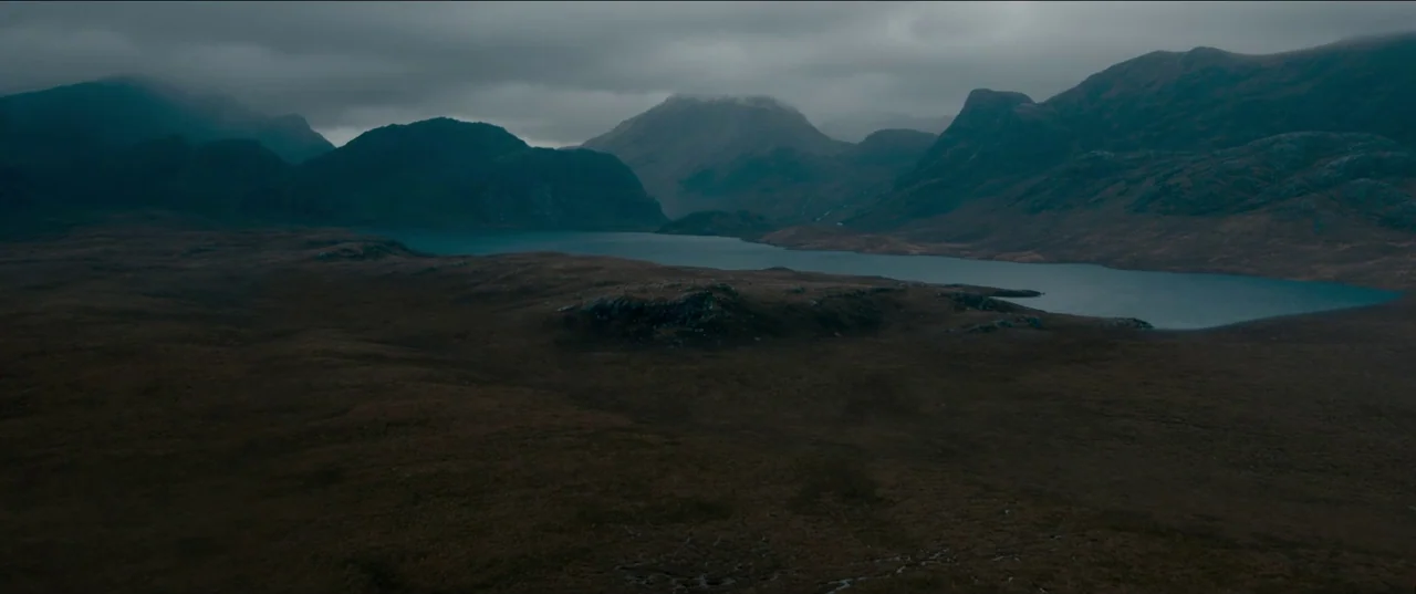 Out of Darkness (2022) movie still: establishing — A vast, rugged highland valley contains a calm dark lake surrounded by rocky ridges. Low-hanging…