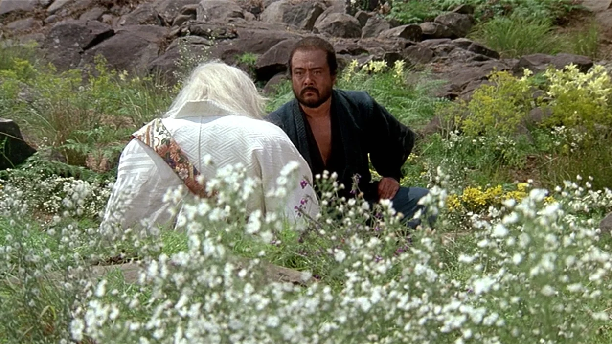Ran (1985) movie still: medium — Two men kneel in a field filled with white wildflowers and scattered rocks. The man in dark clothing looks forward…