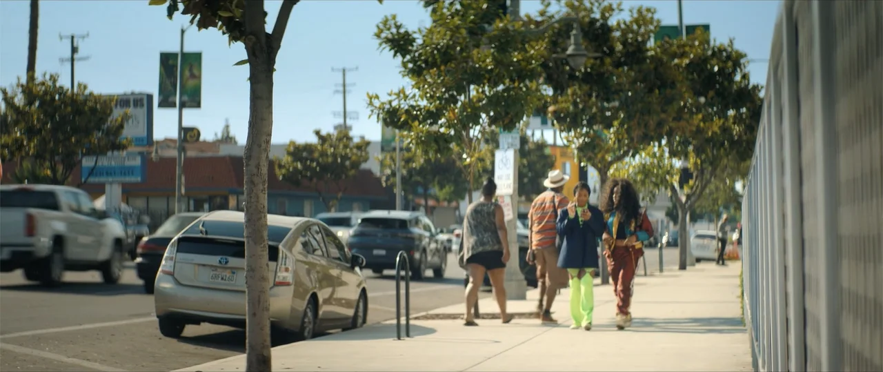 One of Them Days (2025) movie still: fullbody — A sunny urban sidewalk lined with trees and street furniture runs parallel to traffic. Three…