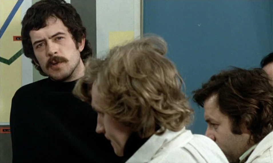 Tout Va Bien (1972) movie still: medium — An interior office space features a wall diagram with lines and text in view. Three men stand together…