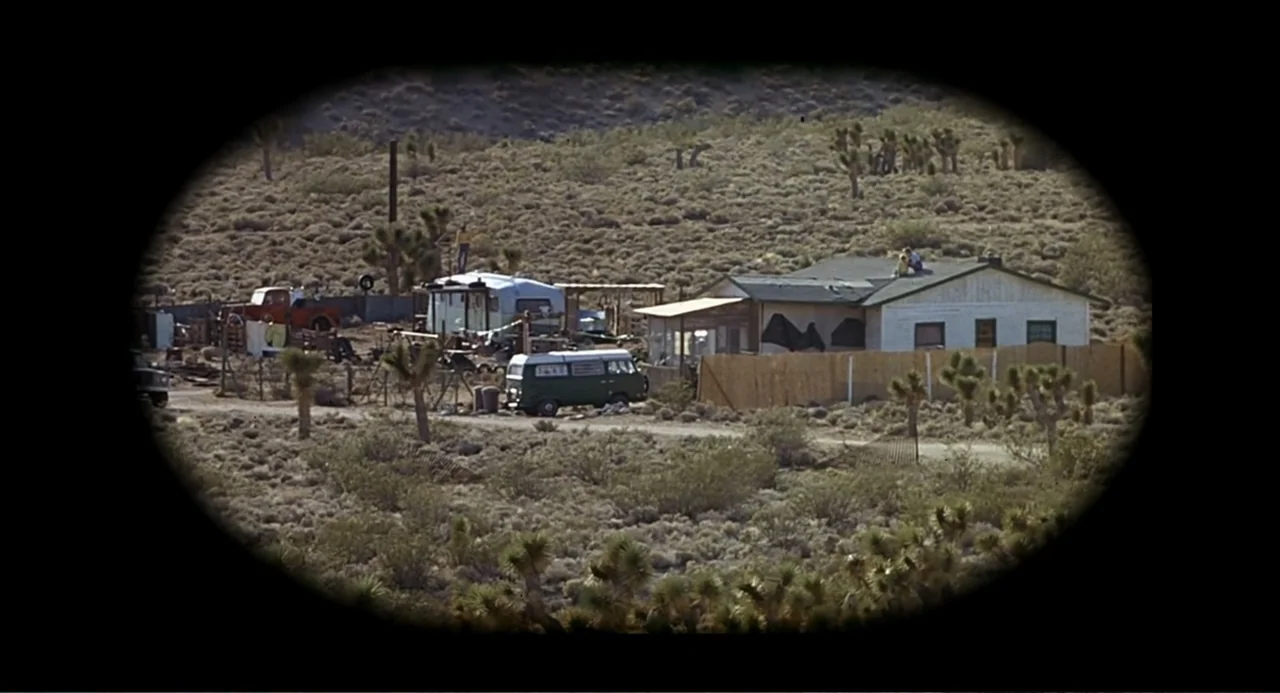 Tremors (1990) movie still: wide — A rural desert property features a small white house, a trailer, and parked vans. Joshua trees dot the…