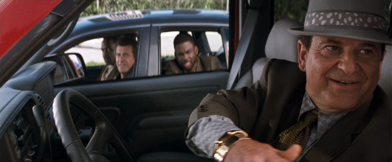 Lethal Weapon 4 (1998) movie still: medium — The interior of a vehicle shows the driver in the foreground and passengers visible through the window.…