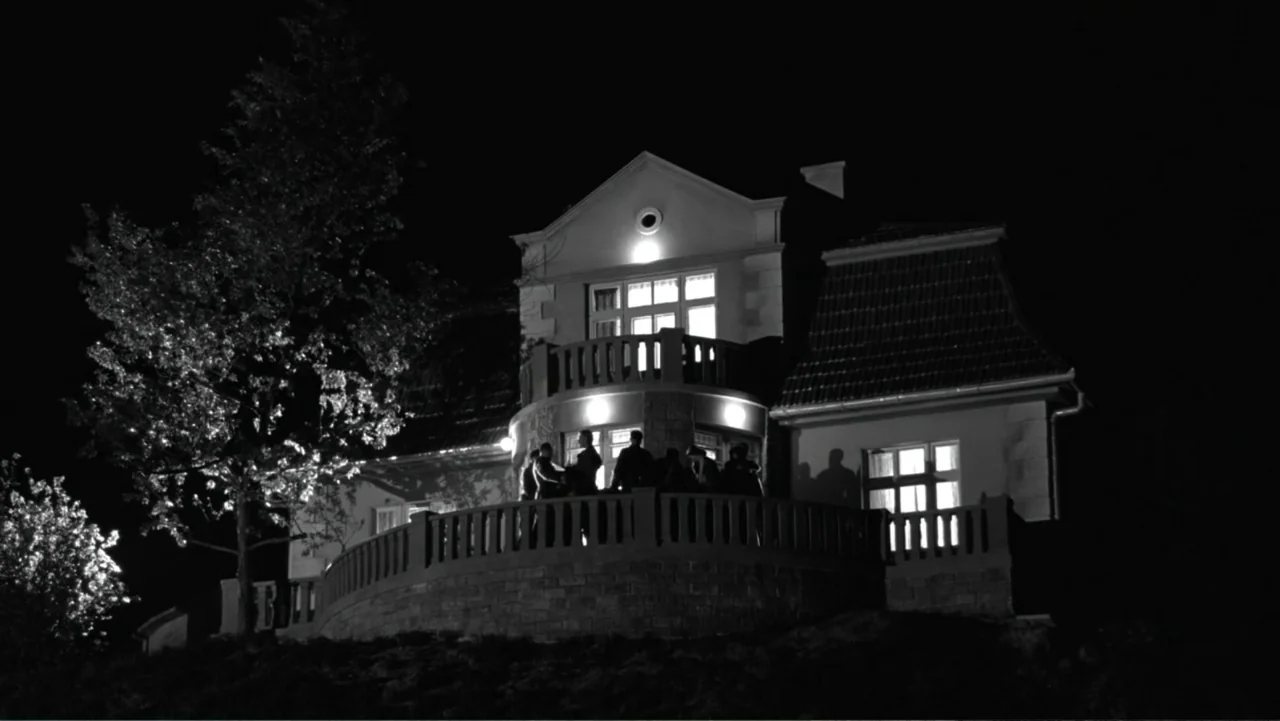 Schindler's List (1993) movie still: wide — A dark night exterior features a large multi-story stone house with a curved balcony. A small group of…