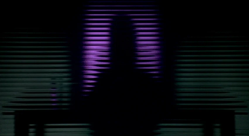 Gray’s Anatomy (1996) movie still: medium — A dark space features a figure sitting before a window covered by horizontal blinds. Purple light…