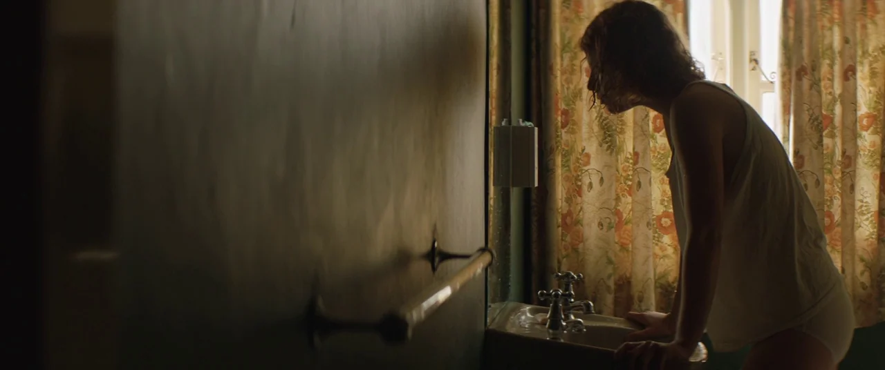 Beast (2017) movie still: medium — A dark bathroom contains a small porcelain sink positioned against a window with floral curtains. A woman in a…