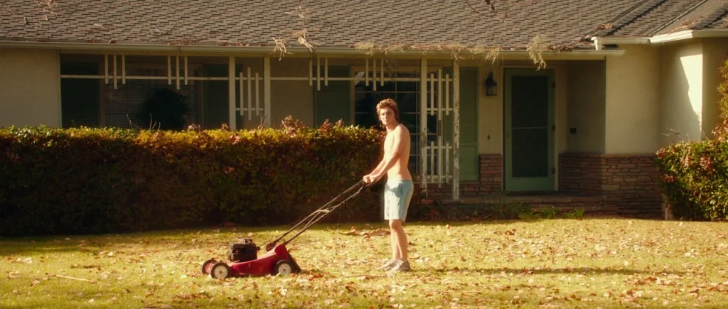 White Bird in a Blizzard (2014) movie still: fullbody — A single-story suburban house sits in the background behind a dry lawn. A young man pushes a…