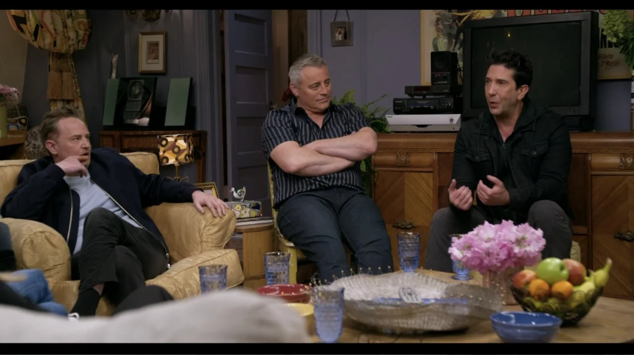 Friends: The Reunion (2021) movie still: medium — A furnished living room features a yellow armchair and a wooden sideboard cabinet. Three…