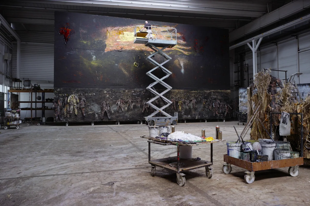 Anselm (2023) movie still: wide — A cavernous industrial art studio contains a massive dark painting on the back wall. An artist on a scissor lift…