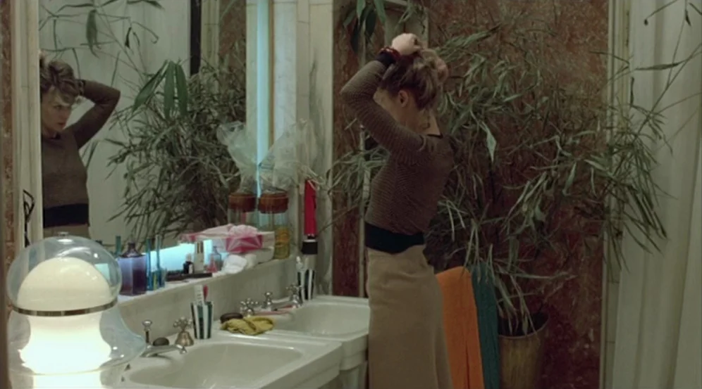 Don’t Look Now (1973) movie still: medium — A tiled bathroom features a double vanity and a tall, leafy potted plant. A young woman stands at the…