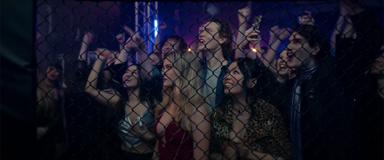 Love Me Love Me (2026) movie still: medium — A crowded nightclub interior features guests gathered behind a black wire chain link fence. Several…