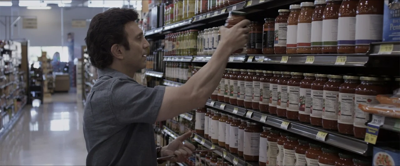 Heavenly Deposit (2019) movie still: medium — A well-stocked supermarket aisle features rows of glass jars of sauce on metal shelves. A man reaches…