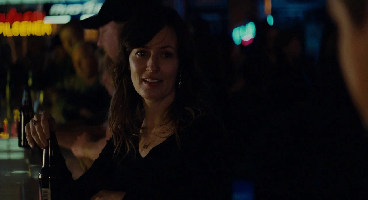 Promised Land (2012) movie still: medium — A dark, crowded establishment is filled with glowing neon signs and patrons at counters. A woman sits at…