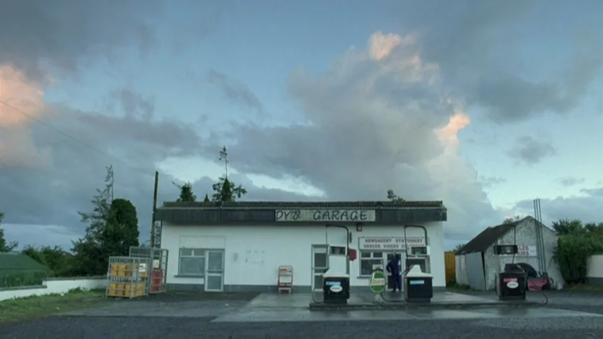 Garage (2007) movie still: wide — A single-story white building labeled DY'S GARAGE sits in a remote, flat area. Two fuel pumps stand in the…