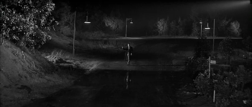 Compulsion (1959) movie still: wide — A rural asphalt road at night features scattered streetlamps along the path. A man in a dark coat stands in…