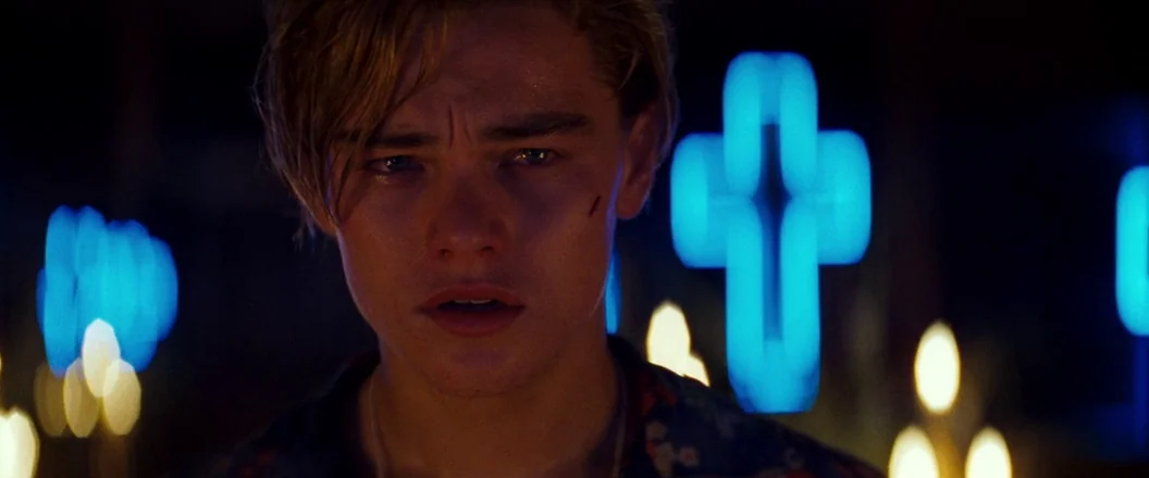 Romeo + Juliet (1996) movie still: closeup — A dimly lit religious interior space features blurred blue neon crosses and candle flames. A young man…