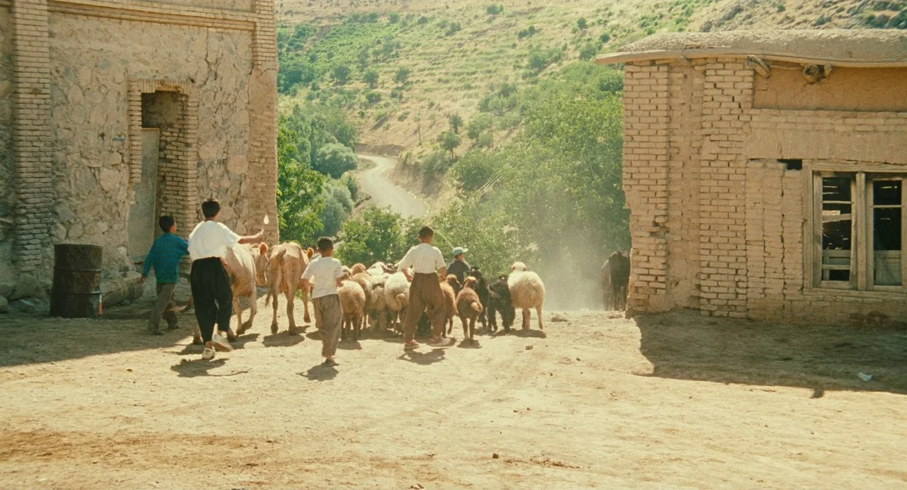 The Wind Will Carry Us (1999) movie still: wide — A rural settlement features two weathered brick buildings flanking a central dirt path. Several…