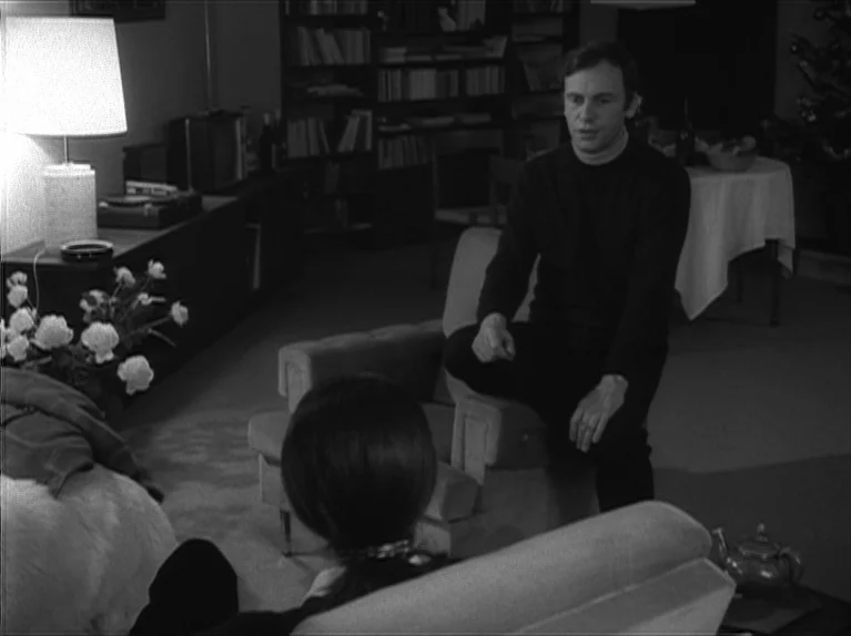 My Night At Maud’s (Ma Nuit Chez Maud) (1969) movie still: medium — A furnished interior space with a bookshelf, table lamp, and armchair. A man…