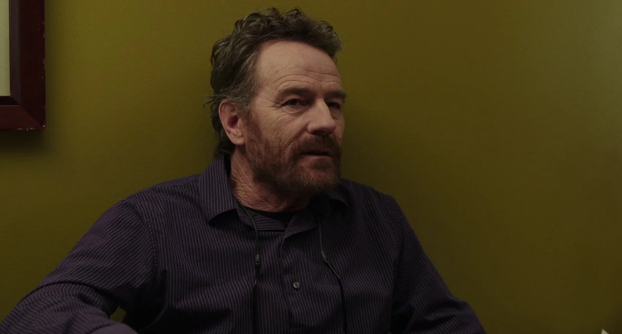 Last Flag Flying (2017) movie still: medium — A man sits against an olive green wall in what appears to be a waiting room. The man is positioned in…