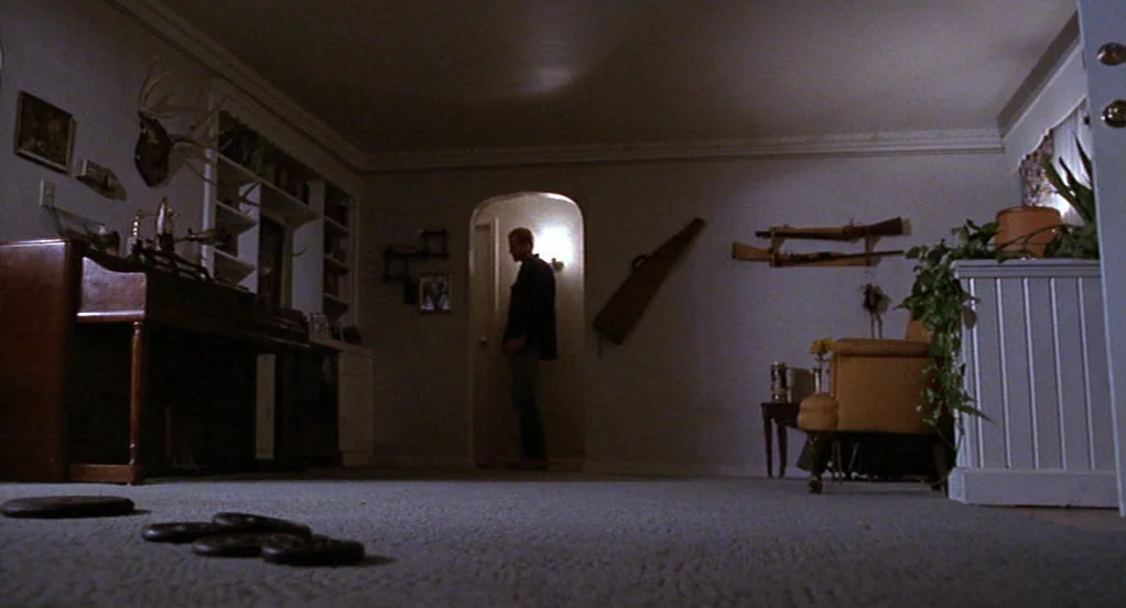 The Indian Runner (1991) movie still: wide — A dimly lit room with carpeted floors displays hunting trophies and antique rifles on walls. A man…