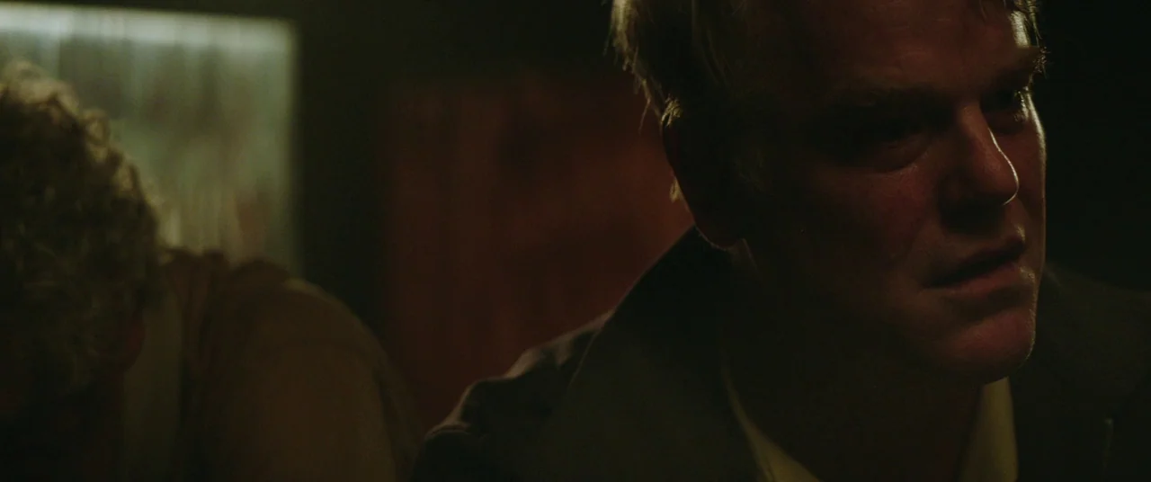 God’s Pocket (2014) movie still: closeup — A dark, minimally lit room contains two figures in low-light conditions. A man's face is illuminated…