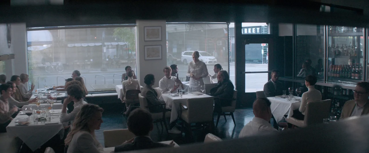 Pig (2021) movie still: wide — A modern restaurant features multiple tables with white tablecloths and patrons seated throughout. A waitress stands…