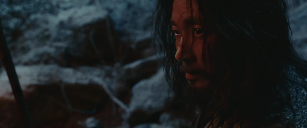 Along with the Gods: The Last 49 Days (2018) movie still: closeup — A dark outdoor space filled with jagged rocks and shadowy textures. A man with…