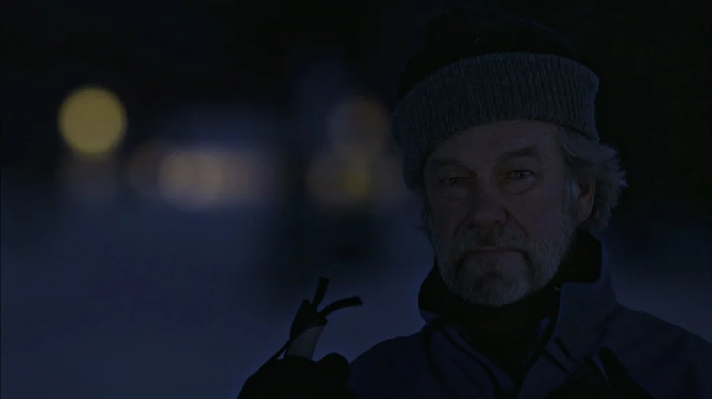 Away From Her (2006) movie still: closeup — A dark, out-of-focus outdoor environment at night with distant, blurry yellow light sources. An older…