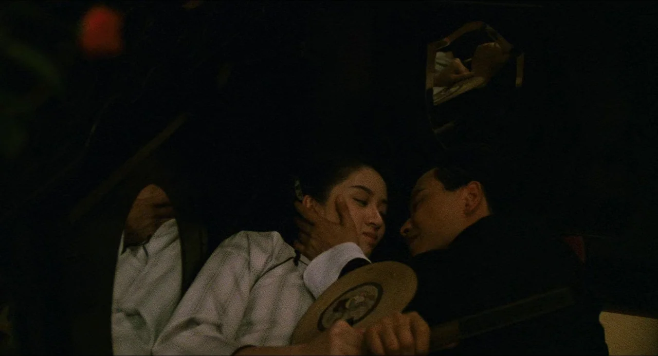 Rouge (1987) movie still: medium — A dimly lit interior space with dark walls and partial reflections in glass surfaces. A man and a woman in formal…