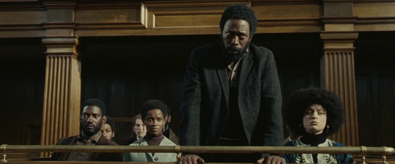 Mangrove (2020) movie still: medium — A group of people sits in the wooden pews of a courtroom. A man stands in the foreground gripping the…