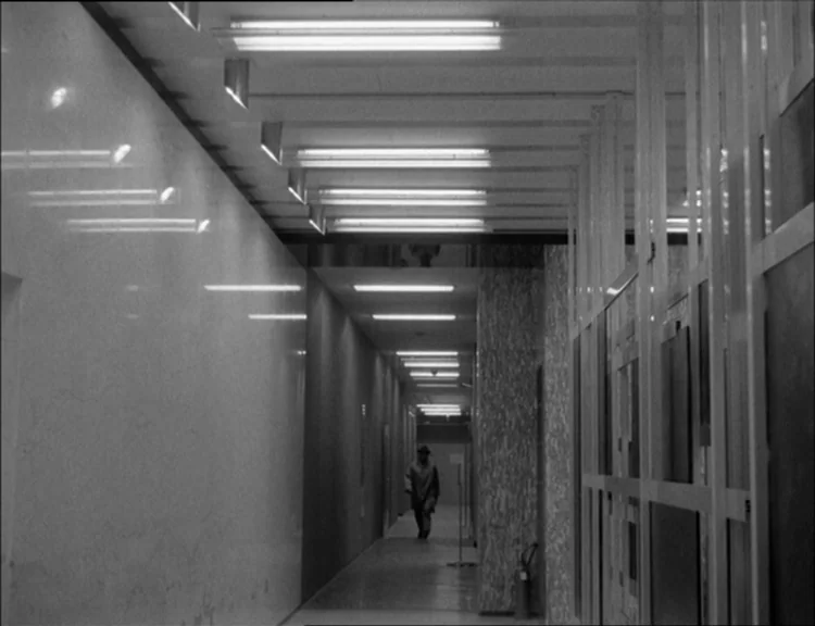 Alphaville (1965) movie still: wide — A long minimalist office hallway with polished light-colored marble walls and geometric ceiling lights. A…