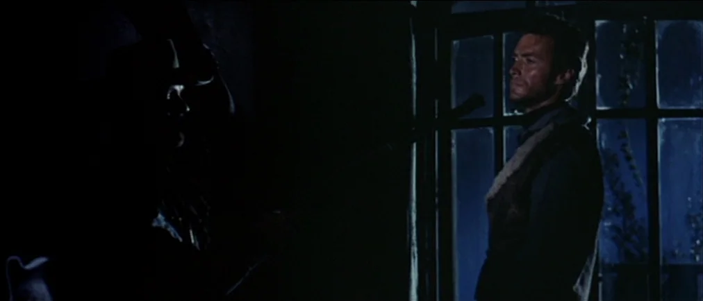 Fistful Of Dollars (1964) movie still: medium — A dark, sparse interior room features a window with a grid frame at night. One man stands visible in…