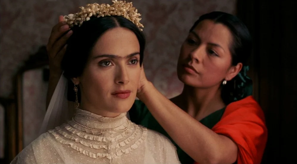 Frida (2002) movie still: medium — An interior bedroom space with faded floral patterned wallpaper serves as the backdrop. A woman standing behind…