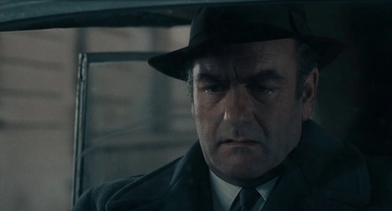 The Army of Shadows (1969) movie still: closeup — The interior of a car features dark upholstered surfaces and a window frame visible. A man sits in…